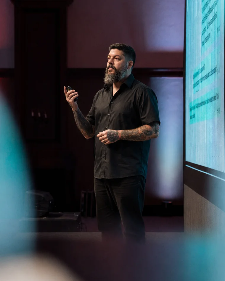 Rodrigo Bondioli on stage in a black shirt, tattooed arms, speaking with a clicker in hand next to a large screen.