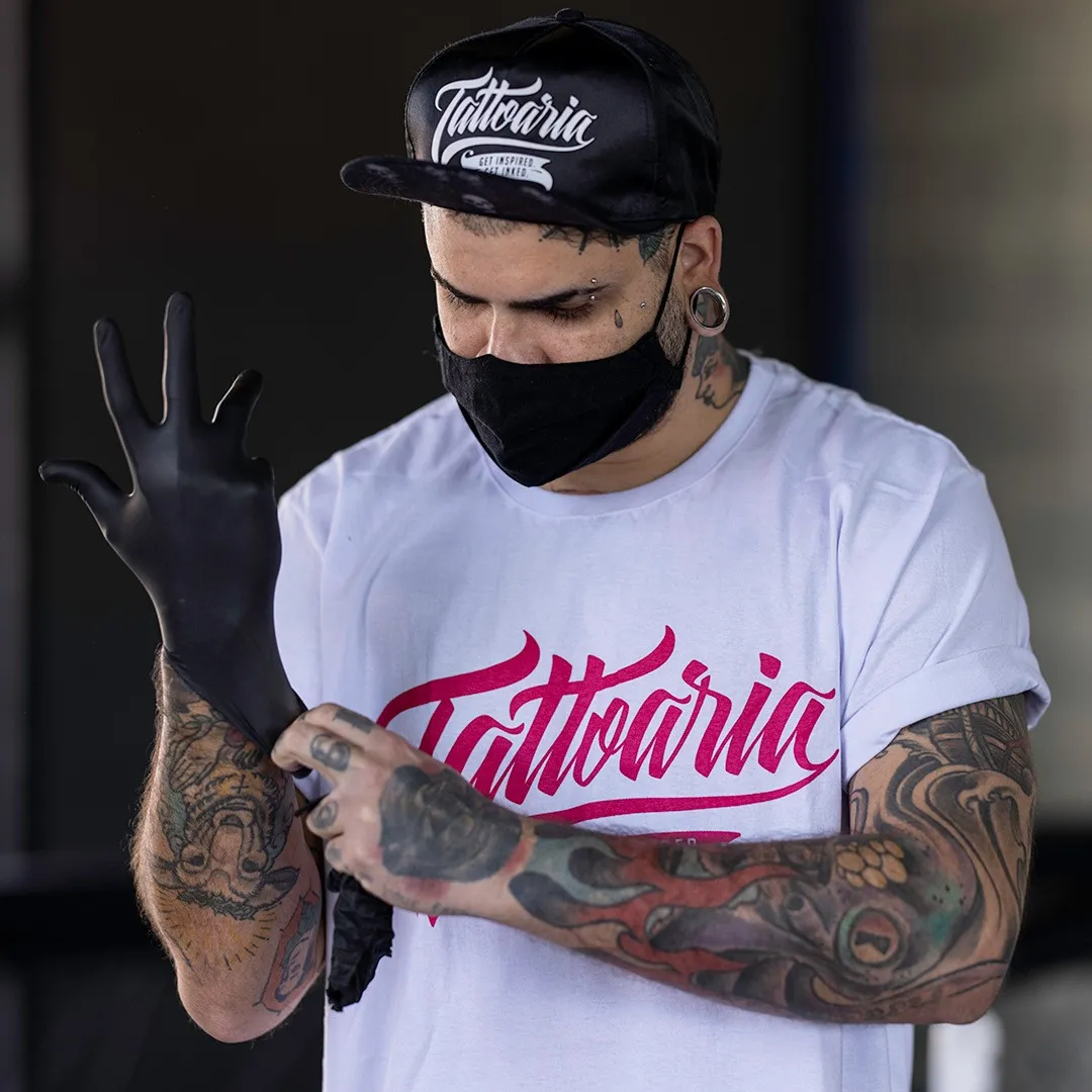 Tatuador convidado de boné e camiseta da Tattoaria calçando a luva preta antes de tatuar.