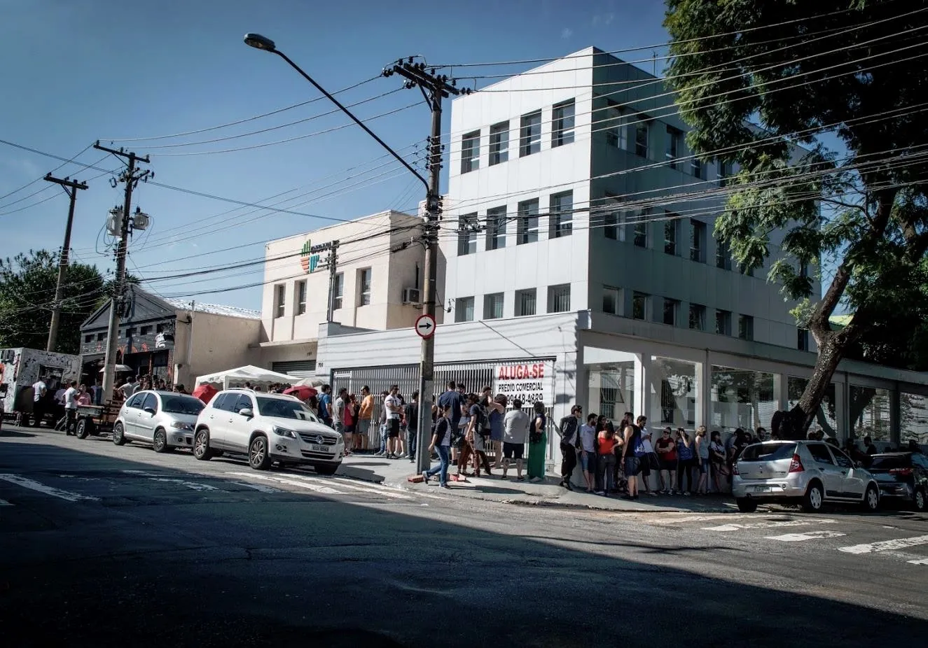 Fila de gente dobrando o quarteirão na porta da Tattoaria num dia de sol, ao longo da calçada.
