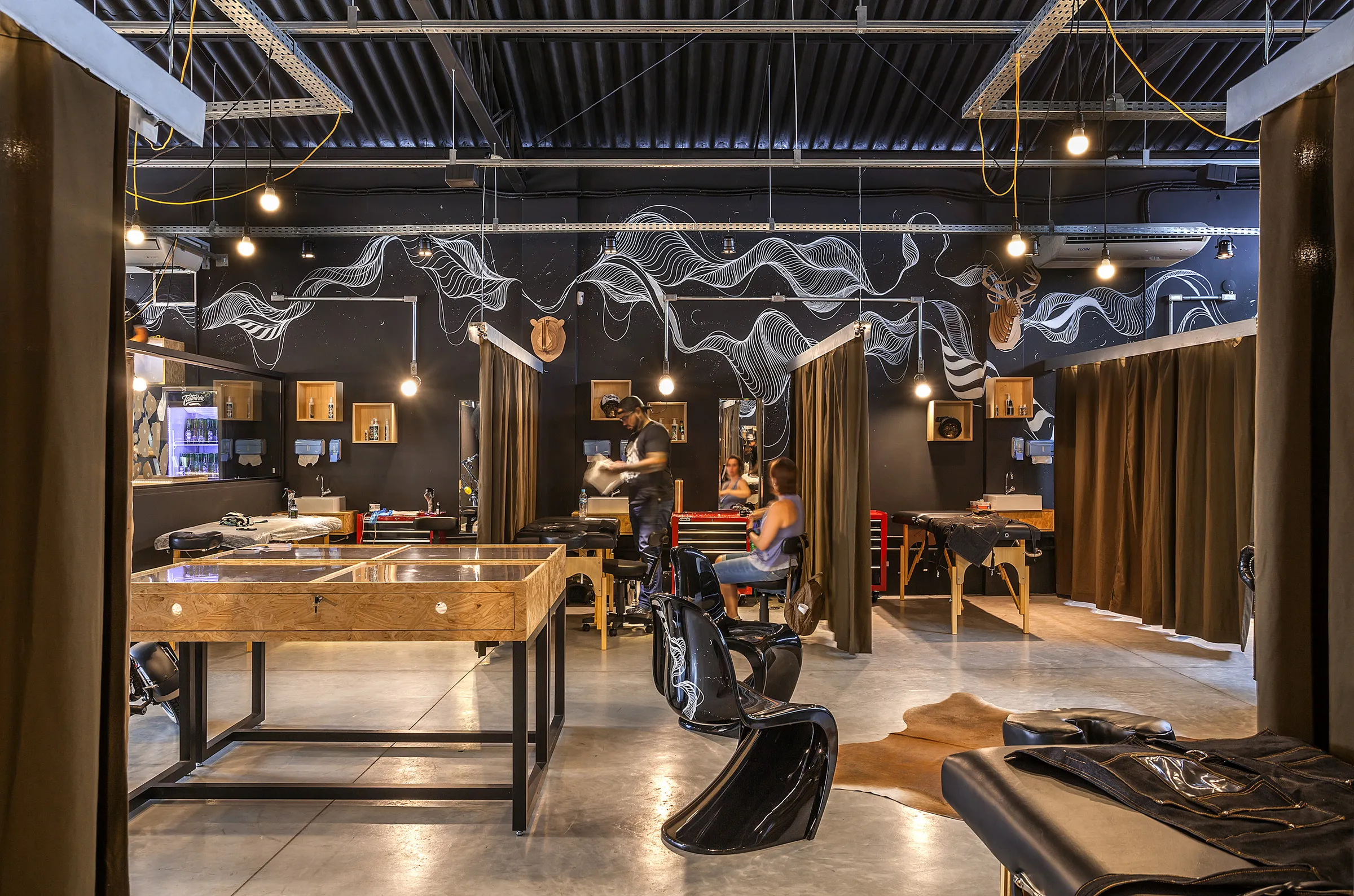 Galpão da Tattoaria: paredes pretas com mural de linhas brancas, baias de tatuagem abertas entre cortinas, vitrine de madeira e um tatuador atendendo uma cliente.