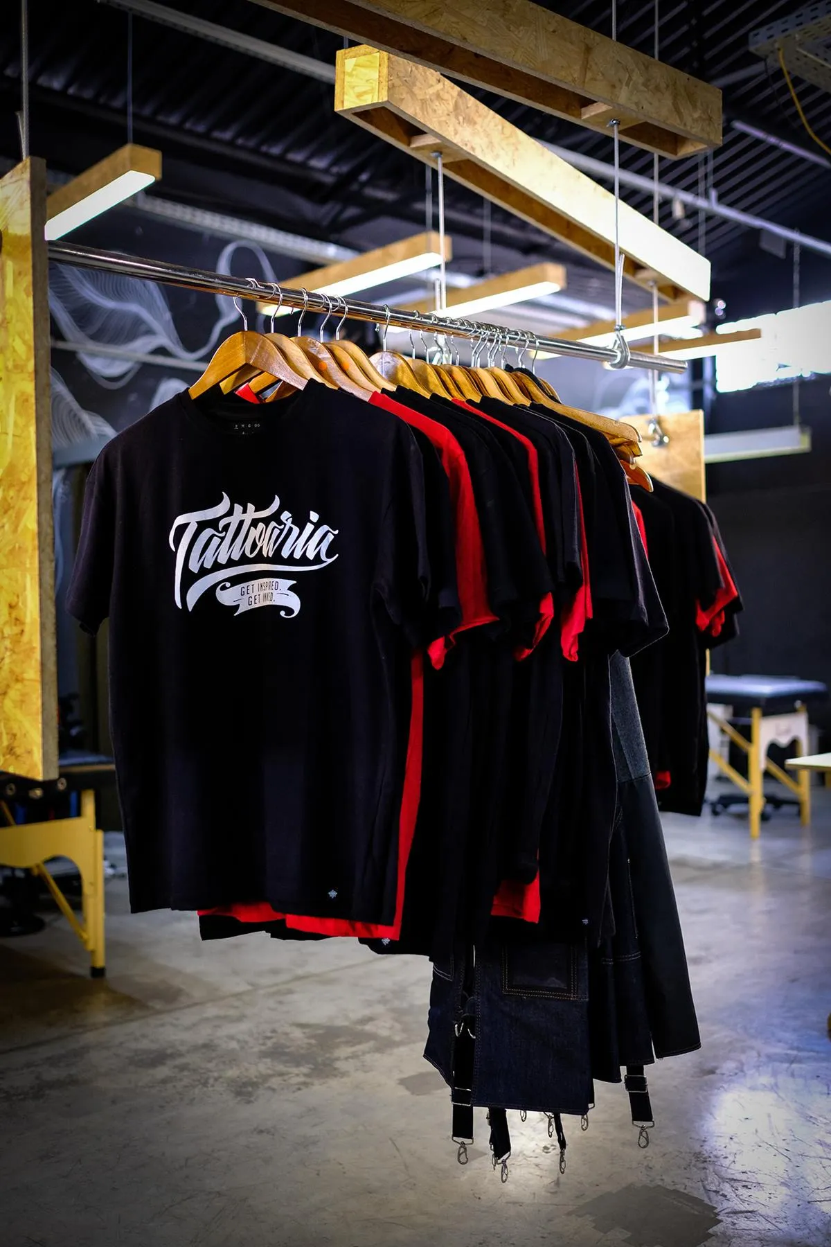 Camisetas pretas e vermelhas da Tattoaria penduradas numa arara de madeira, dentro do galpão.