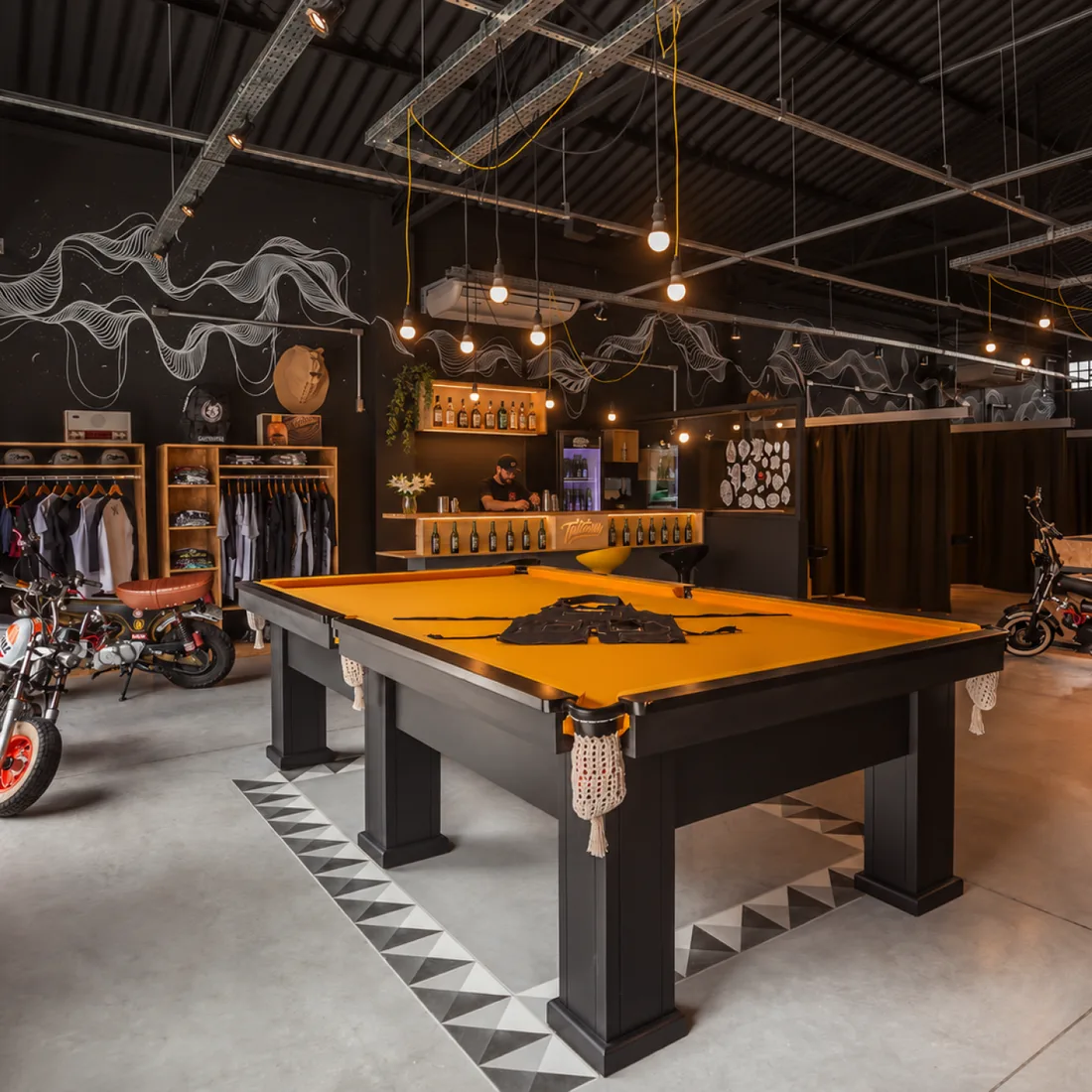 Tattoaria warehouse: a yellow pool table, clothing racks and motorcycles, with black walls and a white line mural